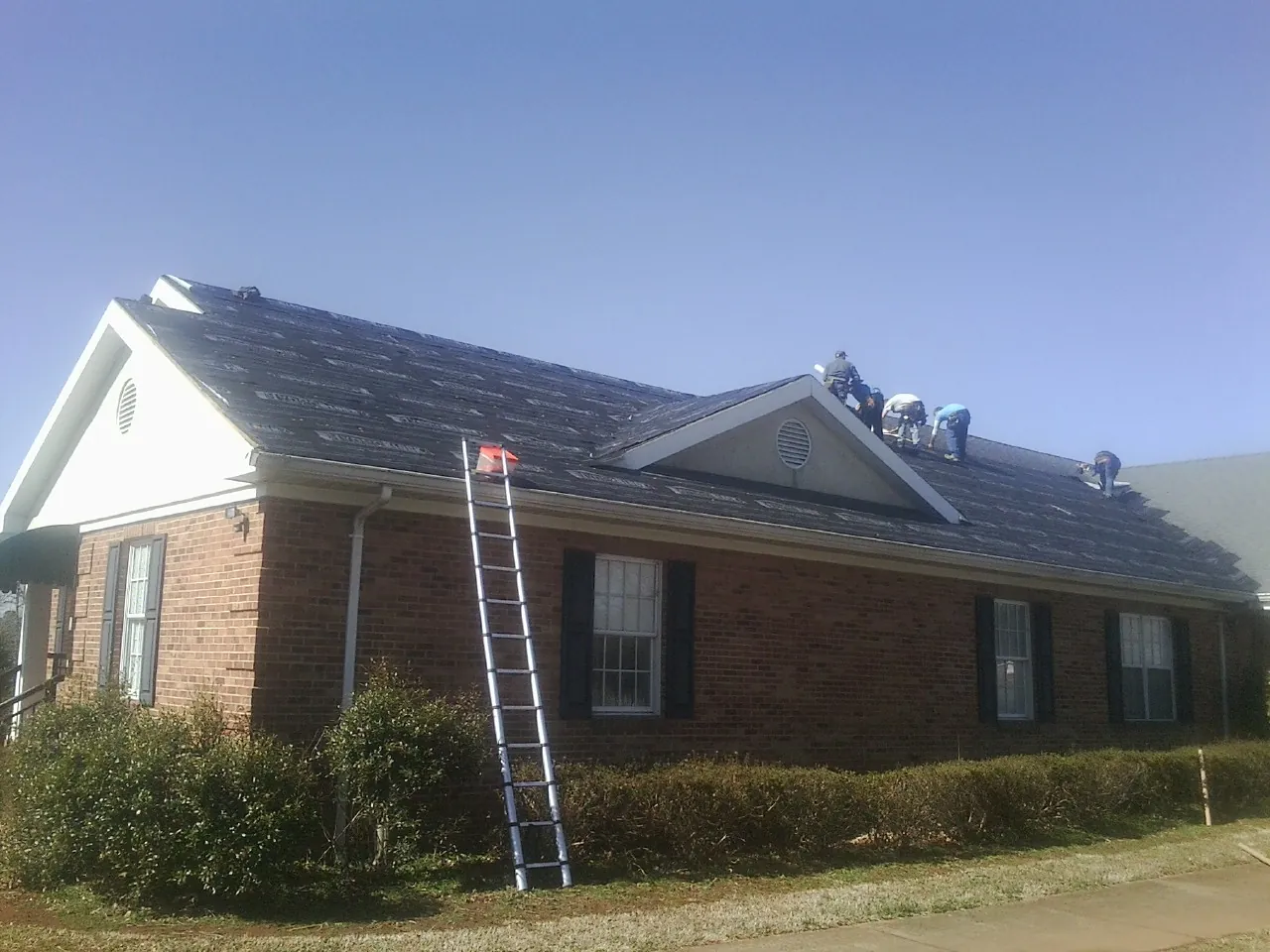 Crew tearing shingles off a brick ranch roof with a ladder set — Metal Roof Repair work by our crew in Johnson Creek