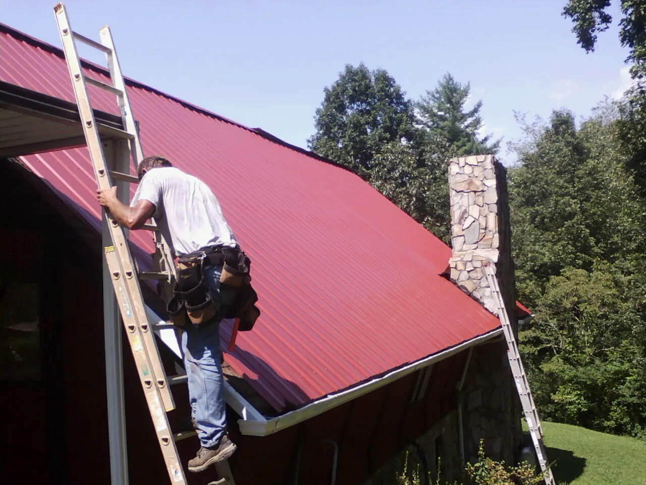 Roofer on a ladder beside a red metal roof and stone chimney — Soffit & Fascia Repair by Roofing Brothers in Johnson Creek